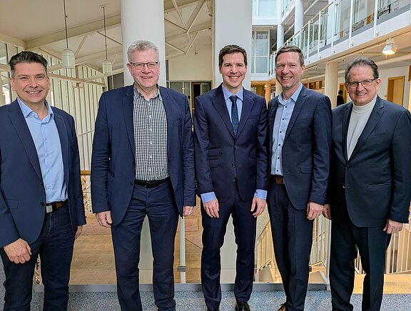 Fünf Männer in Businesskleidung stehen nebeneinander in einem hellen Bürogebäude und lächeln in die Kamera. Gruppenfoto, das ein formelles Geschäftstreffen oder eine Zusammenarbeit vermittelt.