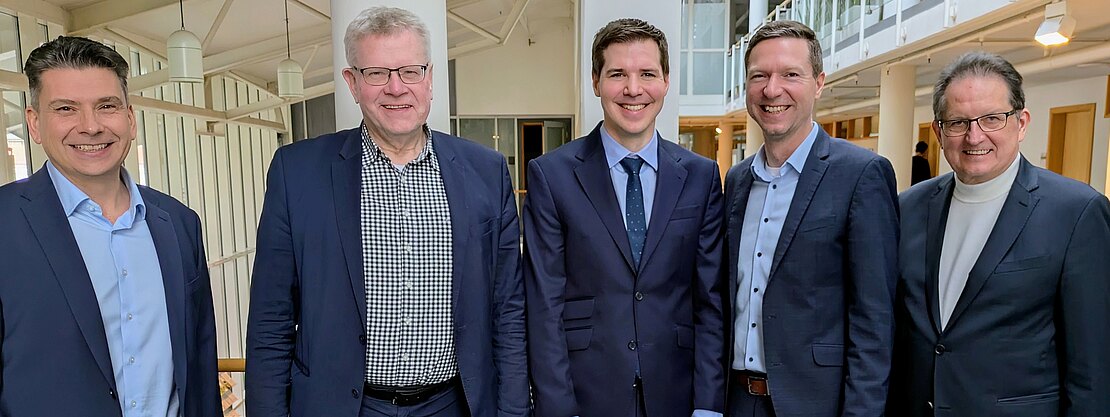 Fünf Männer in Businesskleidung stehen nebeneinander in einem hellen Bürogebäude und lächeln in die Kamera. Gruppenfoto, das ein formelles Geschäftstreffen oder eine Zusammenarbeit vermittelt.
