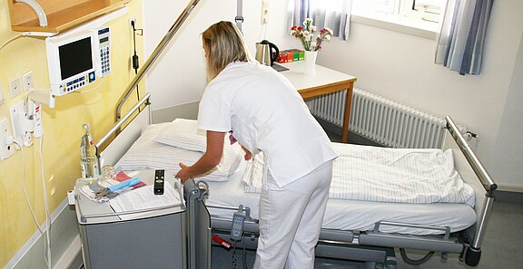 Pflegekraft in weißer Uniform richtet ein Krankenhausbett in einem hellen Patientenzimmer her. Auf dem Nachttisch liegen Pflegeutensilien, daneben medizinische Geräte an der Wand. Szene vermittelt Krankenpflege und Vorbereitung für einen Patienten.