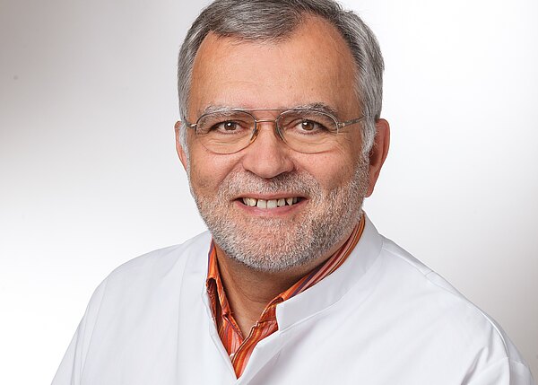 Porträt eines älteren Mannes mit grauen Haaren, Brille und kurzem Bart. Er lächelt freundlich und trägt einen weißen Kittel über einem orangegestreiften Hemd vor neutralem Hintergrund.