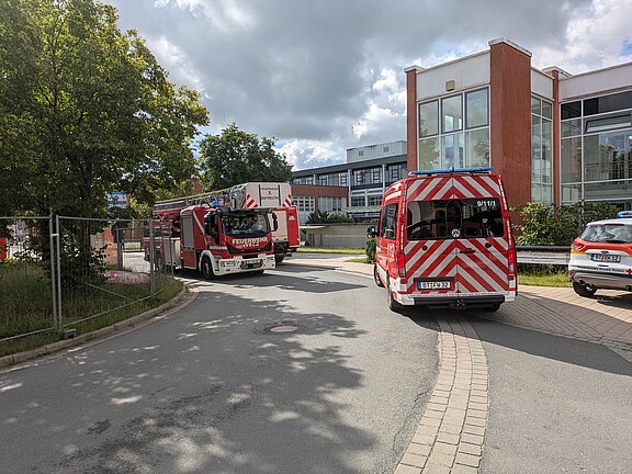 Mehrere Feuerwehrfahrzeuge stehen auf einer Zufahrt vor einem modernen Gebäude mit Glasfassade. Ein Löschfahrzeug und ein Einsatzwagen halten auf der Straße; weitere Fahrzeuge in der Nähe. Keine Flammen oder Rauch sichtbar.