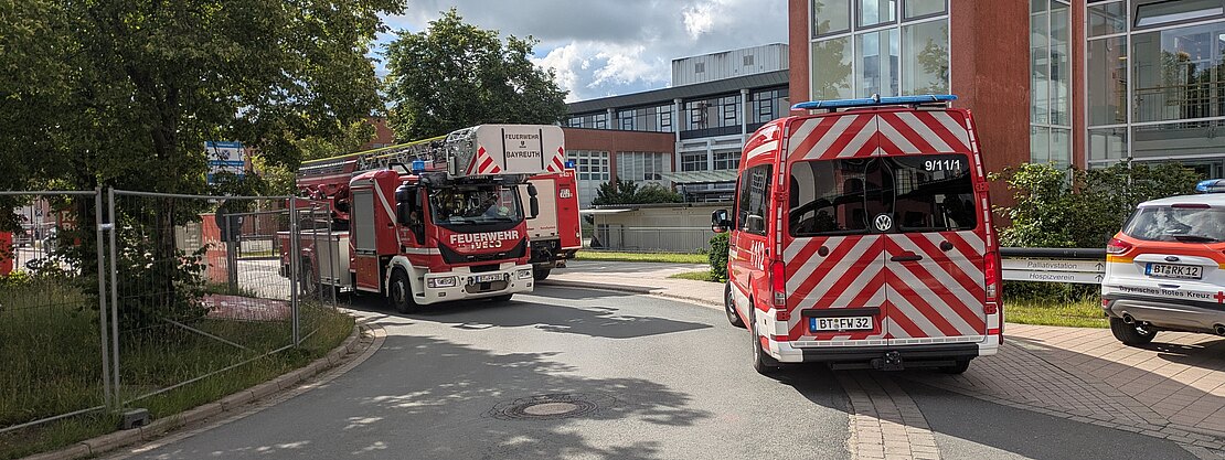 Mehrere Feuerwehrfahrzeuge stehen auf einer Zufahrt vor einem modernen Gebäude mit Glasfassade. Ein Löschfahrzeug und ein Einsatzwagen halten auf der Straße; weitere Fahrzeuge in der Nähe. Keine Flammen oder Rauch sichtbar.