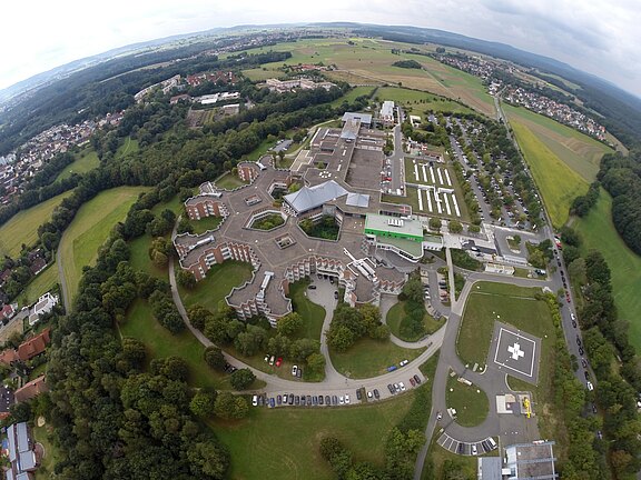 Luftaufnahme eines großen Krankenhausgeländes: mehrere sternförmig angeordnete Gebäudeflügel, zentrale Klinikbauten, lange Parkreihen und ein markierter Hubschrauberlandeplatz. Das Areal liegt zwischen Feldern und Wald, mit angrenzenden Siedlungen im Hintergrund.
