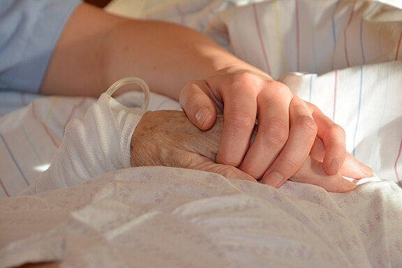 Eine jüngere Hand hält tröstend die Hand einer älteren Person in einem Bett. Ein transparenter Schlauch liegt am Handgelenk der älteren Hand. Die Szene vermittelt Nähe, Unterstützung und Pflege.