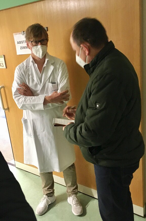 Ein Arzt im weißen Kittel mit Maske spricht im Flur mit einer Person in dunkler Jacke, die in ein Notizbuch schreibt. Beide stehen vor einer Tür mit dem Schild „Abstrichzimmer“.