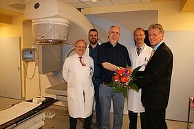 Eine Gruppe von fünf Personen steht in einem modernen medizinischen Raum. Ein Patient in der Mitte hält einen Blumenstrauß, während vier Ärzte um ihn herum stehen. Alle lächeln, was eine positive Atmosphäre vermittelt.
