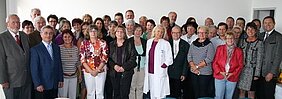 Das Bild zeigt eine große Gruppe von Menschen, die zusammenstehen und in die Kamera schauen. Es sind etwa 50 Personen, darunter Frauen und Männer verschiedenen Alters, einige in Arbeitskleidung, darunter auch eine Person in einem weißen Kittel. Die Szene wirkt freundlich und einladend.
