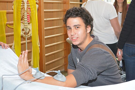 Ein junger Mann sitzt an einem Tisch und hat zwei Elektroden auf seinem Unterarm angebracht. Er lächelt und schaut in die Kamera. Im Hintergrund sind einige Personen und ein Skelettmodell zu sehen.