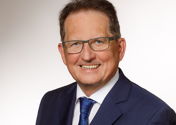 Porträt eines älteren Mannes mit Brille, der freundlich lächelt. Er trägt einen dunkelblauen Anzug, weißes Hemd und eine blaue Krawatte vor hellem, neutralem Hintergrund.