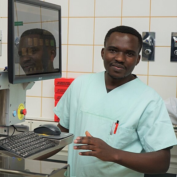 Medizinischer Mitarbeiter in mintgrüner OP-Kleidung steht an einem Computerarbeitsplatz im Krankenhaus und bedient Tastatur und Maus; sterile Kacheln im Hintergrund deuten auf Klinik- oder Laborumgebung hin.