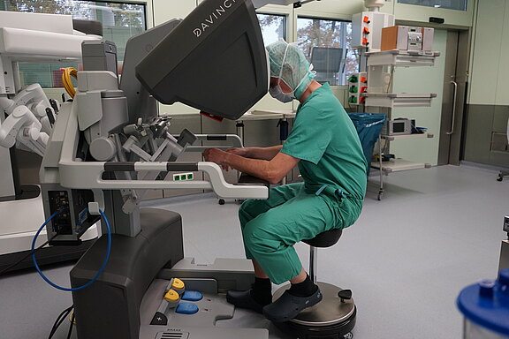 Chirurg in grüner OP-Kleidung bedient in einem Operationssaal eine Roboterchirurgie-Konsole. Kopf in der Sichtkuppel, Hände an den Steuergriffen, Füße auf Pedalen; der Roboter überträgt seine Bewegungen für einen minimalinvasiven Eingriff.