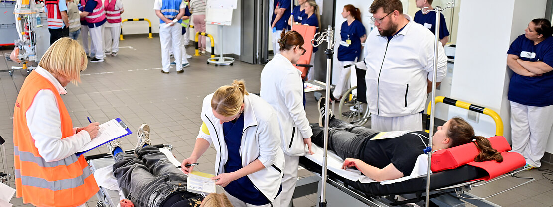 Belebter Krankenhaus-Aufnahmebereich: Medizinisches Personal betreut mehrere liegende Patientinnen und Patienten auf Rolltragen, legt Infusionen an und dokumentiert Befunde. Die Szene wirkt wie eine organisierte Notfall- oder Triage-Situation mit vielen Mitarbeitenden.