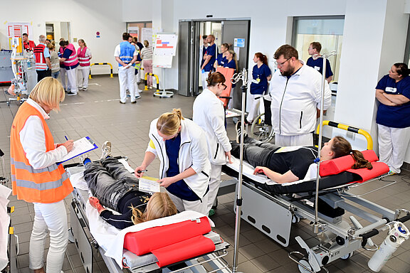 Belebter Krankenhaus-Aufnahmebereich: Medizinisches Personal betreut mehrere liegende Patientinnen und Patienten auf Rolltragen, legt Infusionen an und dokumentiert Befunde. Die Szene wirkt wie eine organisierte Notfall- oder Triage-Situation mit vielen Mitarbeitenden.