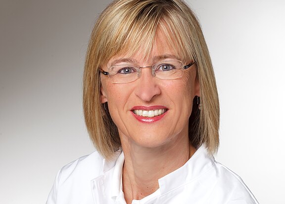 Porträtfoto: Lächelnde erwachsene Person mit schulterlangem blondem Haar und Brille, blickt direkt in die Kamera. Trägt ein weißes Oberteil vor neutralem, hellgrauem Hintergrund.