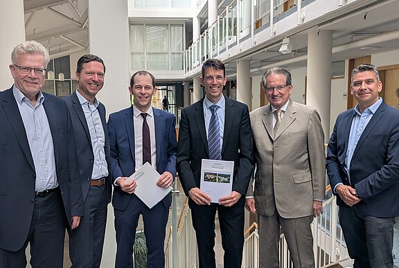 Sechs Personen stehen in einem modernen Bürogebäude, lächeln und halten Dokumente in den Händen. Sie sind formell gekleidet und strahlen eine positive Atmosphäre aus. Im Hintergrund sind helle, offene Räumlichkeiten mit Glaswänden und Treppen zu sehen.