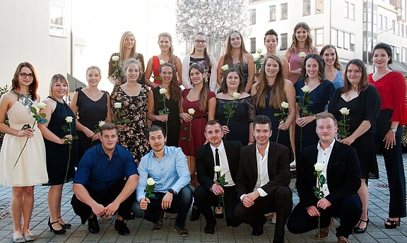 Gruppenfoto: Eine große Gruppe junger Erwachsener in festlicher Kleidung posiert draußen. Viele halten weiße Rosen. Sie lächeln in die Kamera, stehen und knien in Reihen – wirkt wie Abschluss- oder Ehrungsfeier.