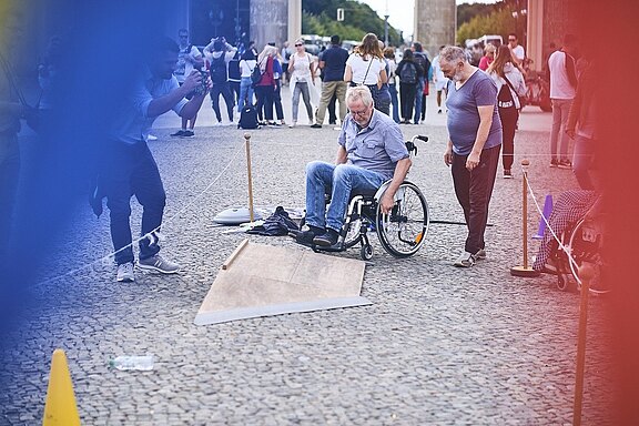 Auf einer belebten Straße testet ein Mann im Rollstuhl eine kleine Rampe eines Hindernisparcours. Neben ihm steht eine Begleitperson, ein Fotograf dokumentiert die Szene. Die Situation vermittelt Barrierefreiheit und Alltagshürden im öffentlichen Raum.