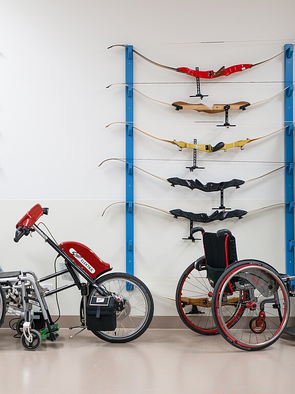 An einer Wand hängen mehrere Sportbögen und Mittelstücke in Halterungen. Davor stehen zwei spezielle Rollstühle, einer sportlich-rot, einer mit Antriebsvorsatz. Die Szene wirkt wie ein Trainingsraum für Para-Bogenschießen.