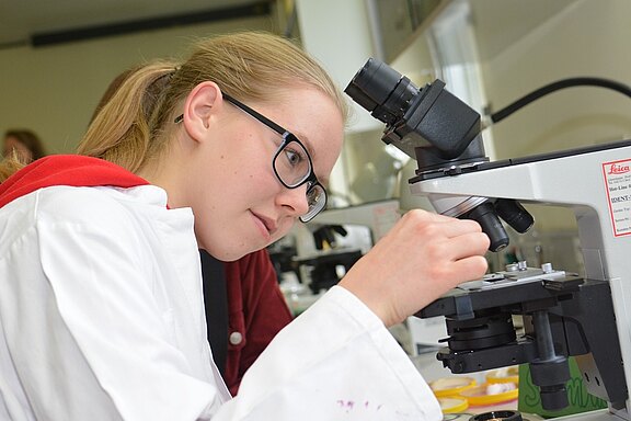 Eine junge Frau mit Brille und Laborkittel schaut konzentriert durch ein Mikroskop. Sie dreht an einem Einstellrad und untersucht einen präparierten Objektträger. Im Hintergrund sind weitere Laborausrüstungen zu erkennen.