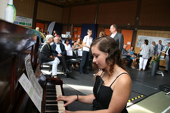 Junge Frau spielt konzentriert Klavier bei einer Veranstaltung in einer Halle. Im Hintergrund sitzen und stehen Zuhörer, einige unterhalten sich. Noten liegen auf dem Klavier. Der Fokus liegt auf der Pianistin, die Musik für das anwesende Publikum spielt.