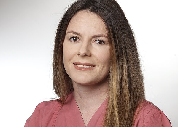 Porträt einer Frau mit schulterlangem braunen Haar, lächelnd, in rosa medizinischem Kasack, vor hellem neutralem Hintergrund, Blick in die Kamera.