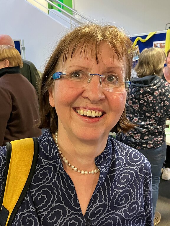 Lächelnde Frau mit Brille, Perlenkette und dunkelblauer, gemusterter Bluse, ein gelber Schulterriemen über der Schulter. Sie schaut direkt in die Kamera. Unscharfer Innenraum mit weiteren Personen im Hintergrund.