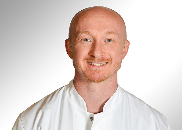 Porträt eines lächelnden, glatzköpfigen Mannes mit kurzem Bart in weißem medizinischem Kittel vor hellem, neutralem Hintergrund; direkter Blick in die Kamera.