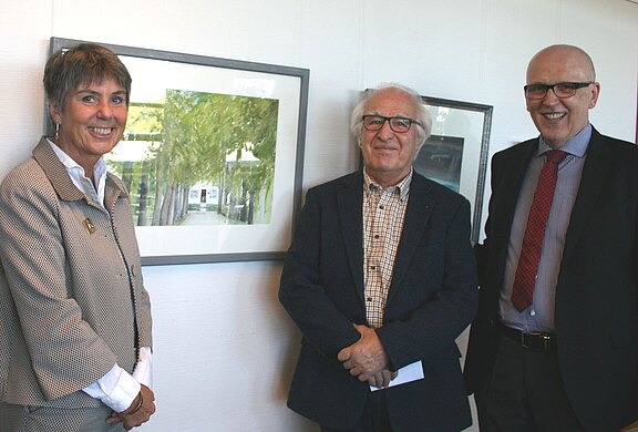 Zwei Männer und eine Frau stehen lächelnd nebeneinander in einer Galerie vor einem gerahmten Bild mit Allee. Sie tragen formelle Kleidung; der Mann in der Mitte hält Papier.