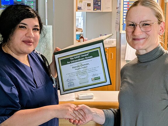 Zwei Frauen im Krankenhaus: Eine Pflegekraft in dunkelblauer Kleidung hält eine Auszeichnungsurkunde und schüttelt der anderen die Hand. Die Szene zeigt die Übergabe eines DAISY-Awards für außergewöhnliche Pflege.