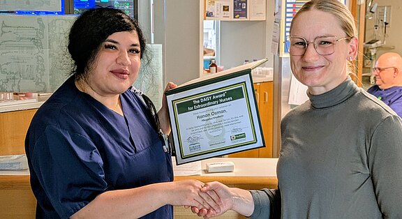 Zwei Frauen im Krankenhaus: Eine Pflegekraft in dunkelblauer Kleidung hält eine Auszeichnungsurkunde und schüttelt der anderen die Hand. Die Szene zeigt die Übergabe eines DAISY-Awards für außergewöhnliche Pflege.