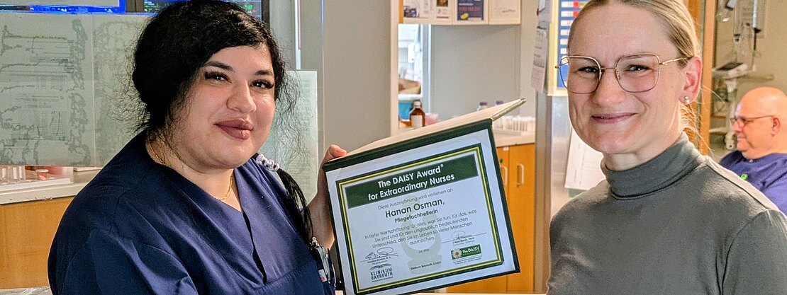 Zwei Frauen im Krankenhaus: Eine Pflegekraft in dunkelblauer Kleidung hält eine Auszeichnungsurkunde und schüttelt der anderen die Hand. Die Szene zeigt die Übergabe eines DAISY-Awards für außergewöhnliche Pflege.