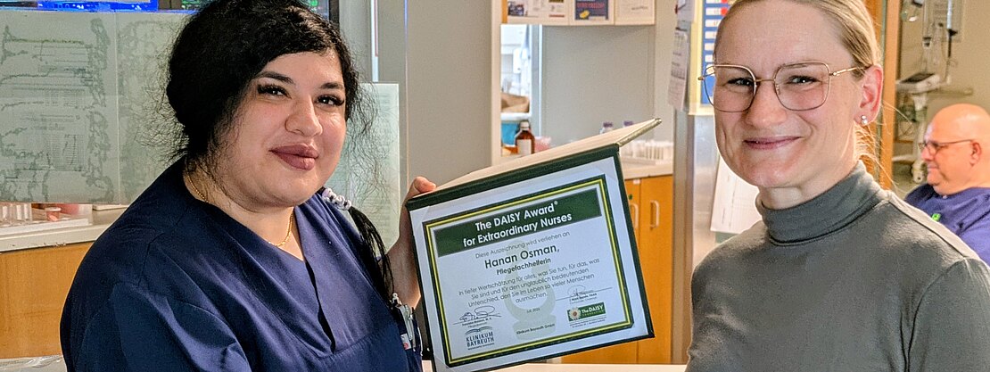 Zwei Frauen im Krankenhaus: Eine Pflegekraft in dunkelblauer Kleidung hält eine Auszeichnungsurkunde und schüttelt der anderen die Hand. Die Szene zeigt die Übergabe eines DAISY-Awards für außergewöhnliche Pflege.