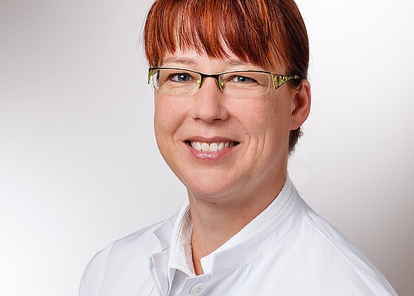 Porträt einer lächelnden Person mit Brille und rötlichen Haaren, die einen weißen Kittel trägt, vor neutralem Hintergrund.