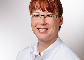 Porträt einer lächelnden Person mit Brille und rötlichen Haaren, die einen weißen Kittel trägt, vor neutralem Hintergrund.