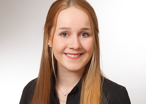 Porträtfoto: junge Frau mit langen, glatten, hellblonden Haaren, lächelt freundlich. Sie trägt eine schwarze Bluse und eine zarte Halskette vor neutralem, hellem Hintergrund.