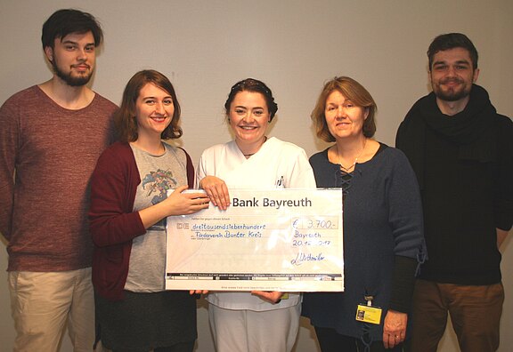 Fünf Menschen stehen nebeneinander und präsentieren einen übergroßen Spendenscheck. In der Mitte trägt eine Person weiße Pflegekleidung. Die Gruppe lächelt in die Kamera; es wirkt wie eine Spendenübergabe zugunsten einer Einrichtung.