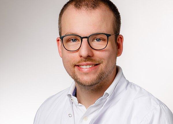 Porträt eines lächelnden Mannes mit Brille, kurzem Haar und leichtem Bart, der einen weißen Kittel trägt. Neutraler Hintergrund.