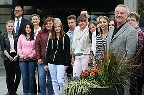 Eine Gruppe von Jugendlichen steht in einem Außenbereich, umgeben von Pflanzen. Einige von ihnen lächeln, während mehrere Erwachsene, darunter ein Mann in einem Anzug, ebenfalls dabei sind. Die Szenerie vermittelt eine freundliche und einladende Atmosphäre.