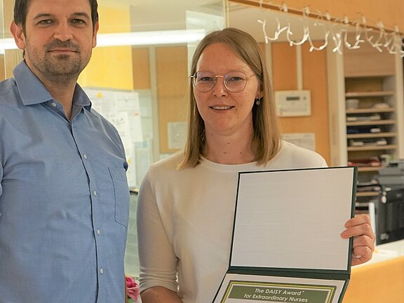 Zwei Personen stehen in einem hellen Empfangsbereich. Die Frau rechts hält eine geöffnete Mappe mit einer Urkunde mit der Aufschrift The DAISY Award for Extraordinary Nurses. Beide blicken lächelnd zur Kamera; es geht um eine Auszeichnung für Pflegekräfte.