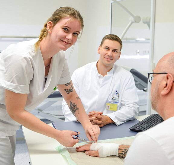 In einem Behandlungsraum versorgt eine Pflegekraft den verbundenen rechten Unterarm eines Patienten am Tisch; sie schneidet Verbandsmaterial. Ein Arzt im weißen Kittel beobachtet die Versorgung. Alle wirken ruhig und konzentriert.