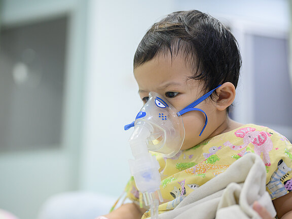 Kleines Kind sitzt in einer Klinik und atmet durch eine Inhalationsmaske (Vernebler). Es trägt einen gelben, bunten Kittel und hält eine Decke. Szene deutet auf Atemtherapie oder Sauerstoffgabe.