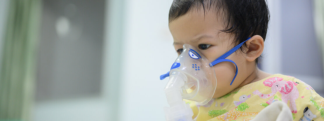Kleines Kind sitzt in einer Klinik und atmet durch eine Inhalationsmaske (Vernebler). Es trägt einen gelben, bunten Kittel und hält eine Decke. Szene deutet auf Atemtherapie oder Sauerstoffgabe.