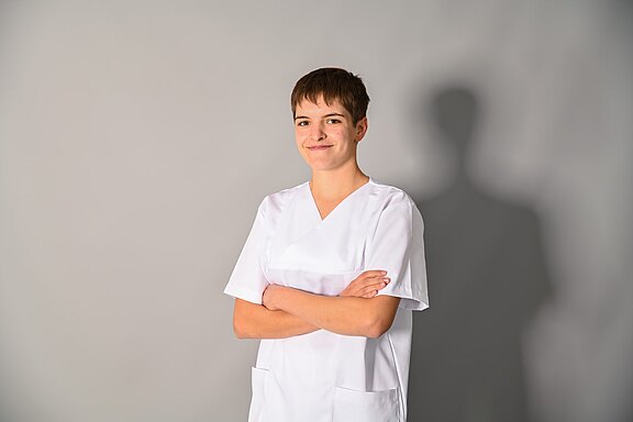 Junge erwachsene Person in weißer Pflegeuniform steht mit verschränkten Armen und lächelt selbstbewusst vor neutralem grauem Hintergrund.