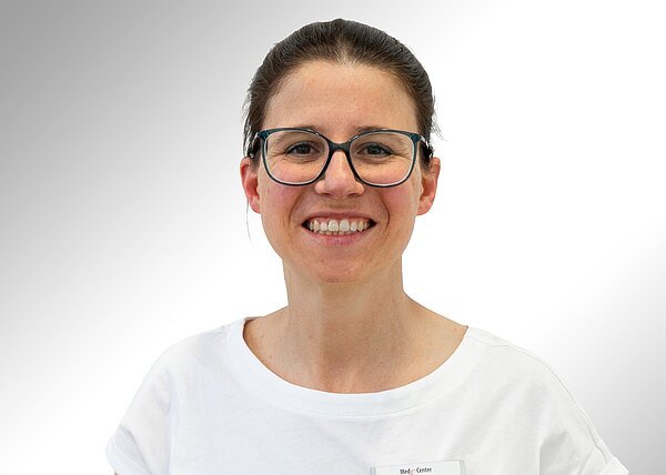 Porträt einer lächelnden Frau mit zusammengebundenen dunklen Haaren, schwarzer Brille und weißem T‑Shirt vor hellem, neutralem Hintergrund.