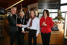 Vier Personen stehen in einem Café und schütteln sich die Hände. Zwei Männer und zwei Frauen sind zu sehen, die freundlich miteinander interagieren. Im Hintergrund sind Schreibtafeln mit Speise- und Getränkenamen sichtbar. Die Szene vermittelt eine Atmosphäre von Zusammenarbeit und positiven Beziehungen.