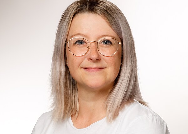 Porträt einer lächelnden Person mit schulterlangem, hellem Haar und runder Brille, im weißen T‑Shirt vor neutralem, hellem Hintergrund. Kopf und Schultern frontal, freundlicher Ausdruck.