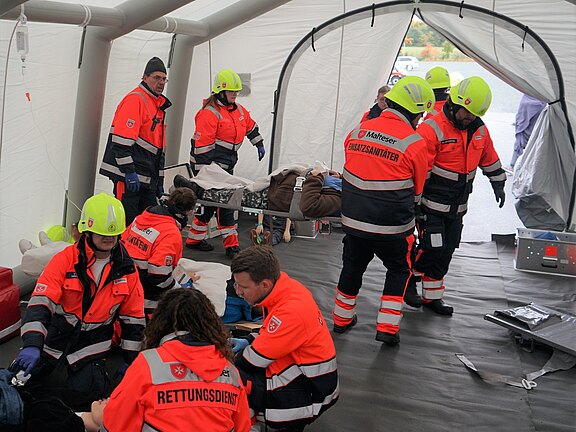 Ein Team von Sanitätern in leuchtend orangefarbenen Uniformen arbeitet in einem Zelt. Einige betreuen Verletzte auf Liegen, während andere medizinische Ausrüstung bereitstellen. Die Szene zeigt eine Notfallsituation mit intensivem Einsatzpersonaleinsatz.