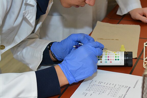 Eine Person in einem weißen Laborkittel führt einen Test durch. Sie trägt blaue Handschuhe und verwendet ein Testbehältnis mit verschiedenen Farbmarkierungen. Neben ihr liegt ein Blatt Papier mit einer Checkliste. Die Szene deutet auf eine wissenschaftliche Analyse hin.