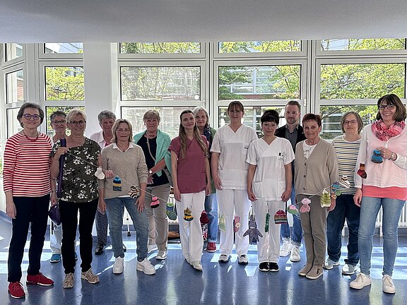 Eine Gruppe von zwölf Personen steht in einem gut beleuchteten Raum mit großen Fenstern. Die Mehrheit trägt lässige Kleidung, einige in Weiß. Sie halten kleine handgefertigte Objekte in den Händen und stehen vor einem Mobile, das aus ähnlichen Objekten besteht.