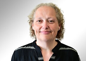 Iris Kammerer Porträt einer Frau mittleren Alters mit kurzen, lockigen Haaren, freundlich lächelnd und in die Kamera blickend. Sie trägt eine schwarze Bluse mit dezentem Muster vor neutralem, hellgrauem Hintergrund.